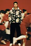 A man in a patterned jacket stands centered as blurred people in red move around him.