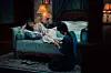 Two people sit beside a bed where another person is lying under a blanket in a dim bedroom.