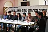 A group of people seated at a table during a press conference with a Korean banner behind them.