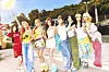 Seven young women stand together outdoors in colorful outfits under bright sunlight.