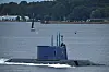 A blue submarine moves through the Kiel Fjord with a forested shoreline in the background.