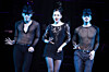 Three performers in black costumes pose under blue lighting on a dark stage.