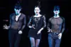 Three performers in black costumes pose under blue lighting on a dark stage.