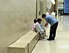 A man leans toward two seated children on a bench in a spacious indoor hallway.