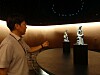 A person stands beside a round display table and points toward two sculptures in a dark gallery.