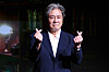 Actor Choi Min-sik poses in a dark suit making finger hearts at a press event.