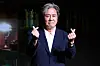 Actor Choi Min-sik poses in a dark suit making finger hearts at a press event.