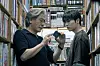Two men stand in a narrow bookstore aisle, studying a small object together.