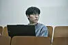A man sits in a lecture hall behind a laptop, looking off to the side.
