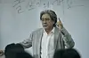 Choi Min-sik stands in a classroom beside a chalkboard with Korean writing, raising one hand near his head.