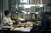 Two actors sit at a desk in a book-lined study with papers, books, and lamps.