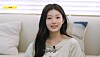 Wonhee of ILLIT speaks on Idol Human Theater in a bright room with a couch and pillows behind her.