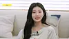 Wonhee of ILLIT speaks on Idol Human Theater in a bright room with a couch and pillows behind her.