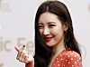Sunmi smiles and makes a finger-heart gesture on a red carpet event backdrop.