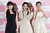 Three women pose in front of a pink event backdrop at a film press conference.