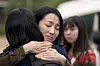 Two women hugging outdoors with another blurred person behind them.
