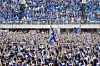 A sports figure in blue is held up by a large cheering crowd in a stadium.