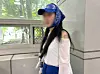 Woman in blue cap and head covering standing beside a window in a tiled corridor.