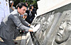 A man in a gray suit and white gloves touches a stone memorial relief beside photographers outdoors.