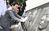 A man in a gray suit and white gloves touches a stone memorial relief beside photographers outdoors.