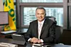 Suited executive seated at a desk in an office with a laptop, window view, and yellow flag.