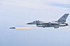A gray KF-16 fighter jet flies in profile while firing a missile with a bright exhaust trail.