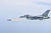 A gray KF-16 fighter jet flies in profile while firing a missile with a bright exhaust trail.