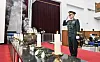 A uniformed military officer salutes beside a casket area decorated with flowers and candles in a memorial hall.