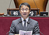 A man in a suit speaks at a podium inside the National Assembly, with microphones and a chair behind him.