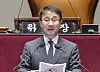 A man in a suit speaks at a podium inside the National Assembly, with microphones and a chair behind him.