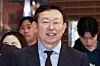 A suited man smiles indoors while people stand behind him in a store or mall entrance.