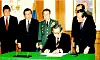 A seated official signs a document at a green-covered table while several suited men stand behind him.