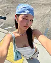 Young woman taking a selfie outdoors on a sunny sidewalk wearing a blue bandana and white top.
