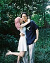 A woman with pink hair hugs a smiling man standing in a green garden.