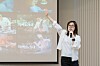 A woman in glasses holds a microphone and gestures next to a projected image collage during a presentation.
