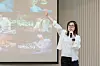 A woman in glasses holds a microphone and gestures next to a projected image collage during a presentation.