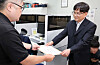 A school official hands a letter to another man in an office, with computer monitors in the background.