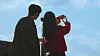 Two silhouetted people stand against a blue sky with clouds, one holding a hand up near their face.