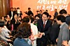A crowd gathers indoors as a man in a suit shakes hands with attendees near a stage with Korean writing.