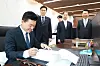 A man in a suit signs a document at a desk while four other suited men stand behind him in an office.