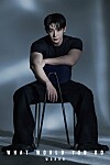Man seated on a chair in a dark studio portrait against a gray background.