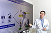 A doctor in a white coat stands beside a large presentation screen in a hospital office.