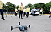 A drone sits on pavement while officials stand in the background at an outdoor demonstration site.