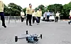 A drone sits on pavement while officials stand in the background at an outdoor demonstration site.