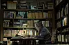 A man sits at a desk in a dimly lit study surrounded by shelves of books and papers.