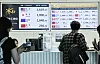 Travelers stand at an airport bank exchange counter with won-dollar rates shown on electronic display boards.