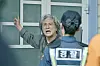 Choi Min-sik in costume gestures beside a police officer outside a green door.