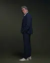 A man in a dark suit stands against a dark studio backdrop wearing white sneakers.