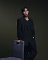 Man in black clothing standing beside a gray chair against a dark green studio background.