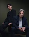 Two men in dark outfits posed together in a dark studio portrait.